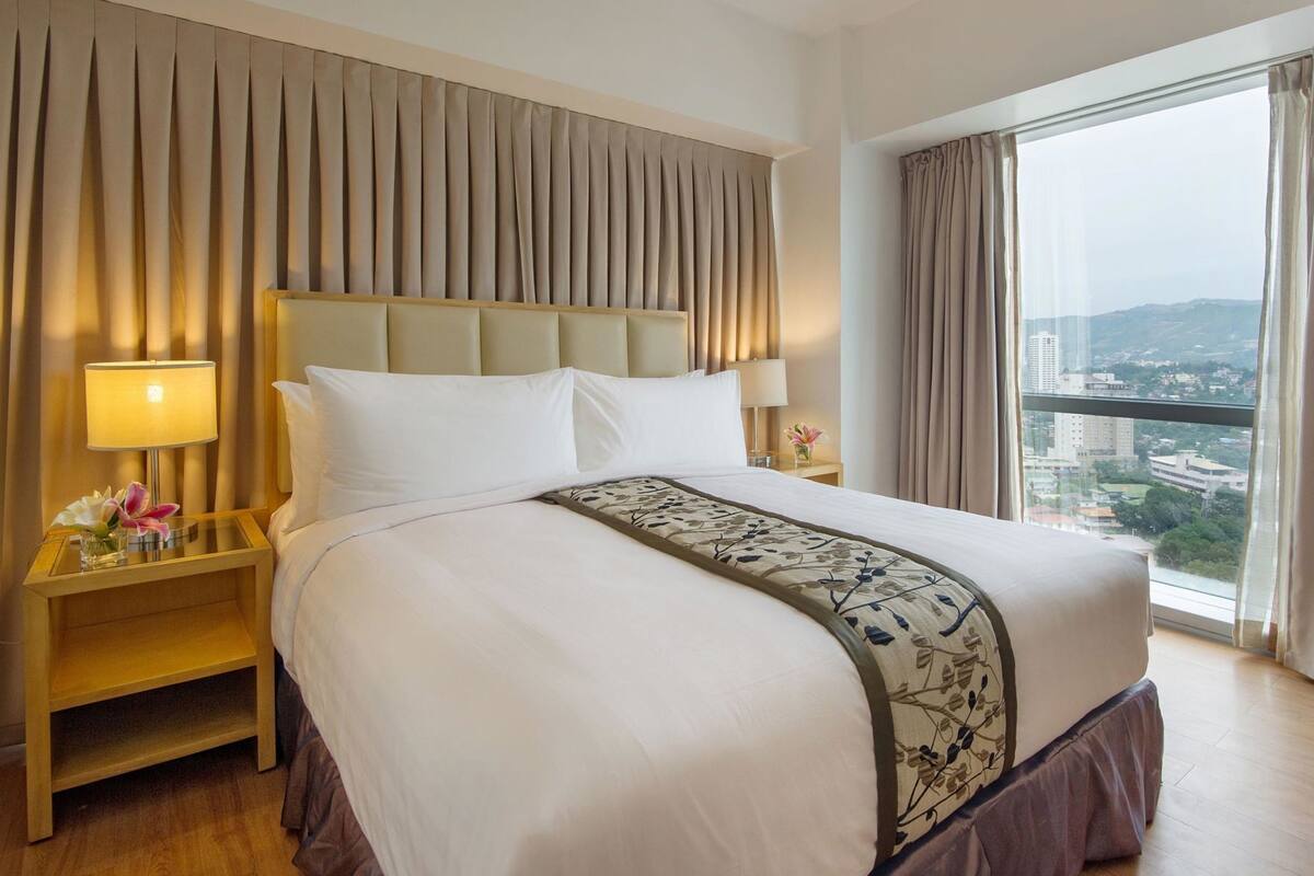 Ein Hotelzimmer mit einem großen Bett, zwei Nachttischen, einer Lampe und Blick auf eine Stadtlandschaft.