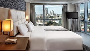 Ein Hotelzimmer mit einem großen Bett, einer Nachttischlampe, einem Telefon, einem Fernseher und Blick auf die Stadt.