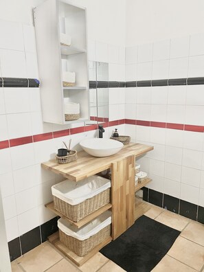 Un baño con un lavamanos blanco, estantes de madera, cestas de mimbre y una alfombra negra.