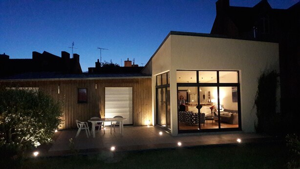Una casa moderna con un'ampia porta vetrata che si apre su un soggiorno, un piccolo patio con tavolo e sedie, e un garage con persiana chiusa.