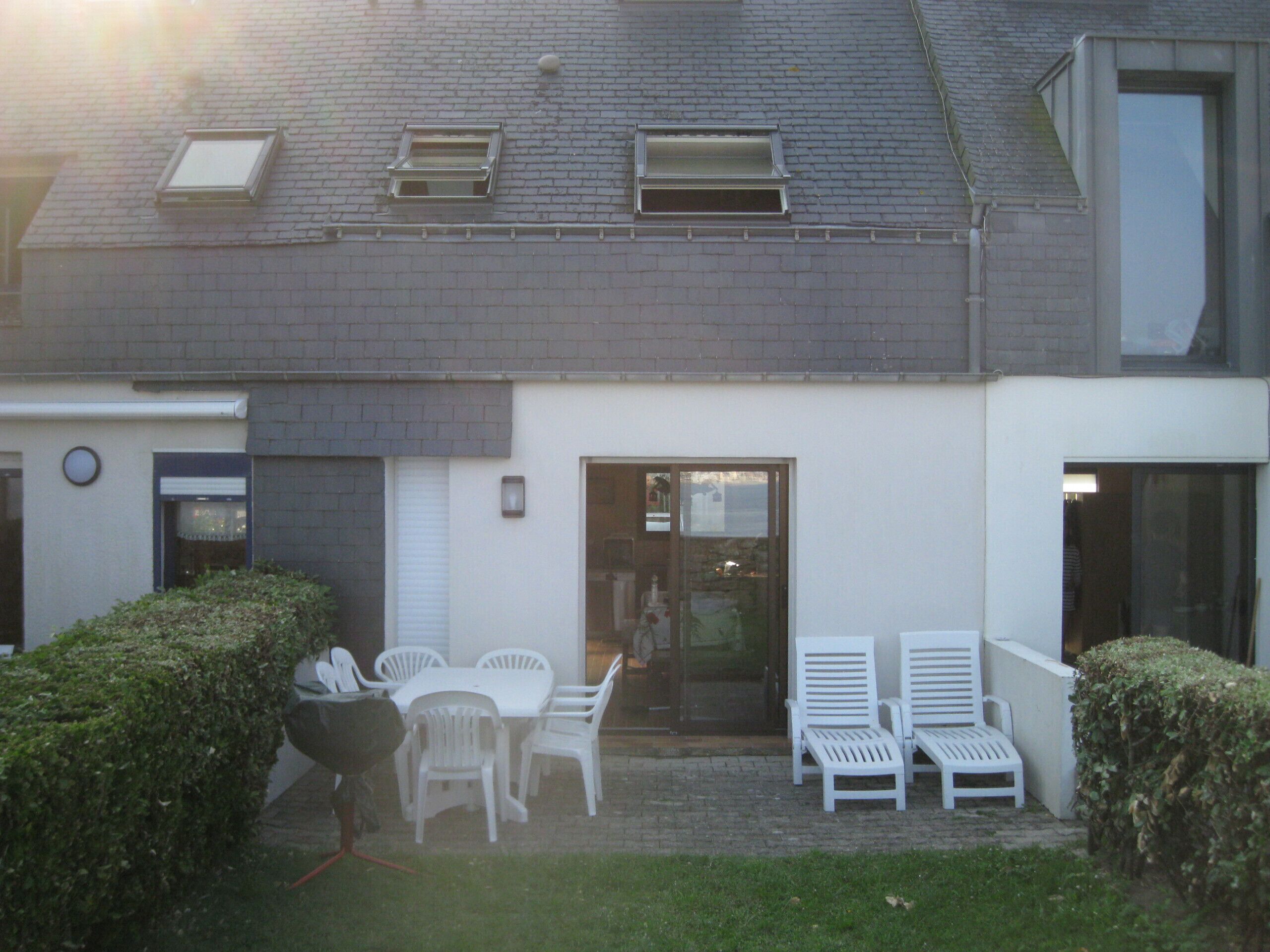 Un bâtiment avec une façade en briques, une porte blanche et une petite fenêtre. On peut voir des chaises blanches et une table à l’extérieur.