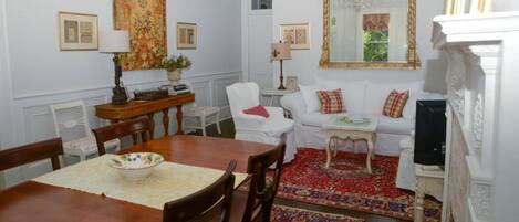 Un coin repas avec une table en bois, des chaises et un saladier. On y trouve un canapé blanc et un tapis à motifs rouges et blancs. Un grand miroir et des tableaux encadrés ornent les murs.
