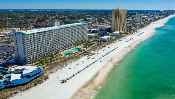 A coastal hotel with a large building, a beach, and a pool area.