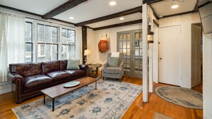 A living room with a brown leather sofa, a wooden coffee table, and a patterned rug. There is a chair with a cushion, a small table with a lamp, and a wall-mounted light fixture.