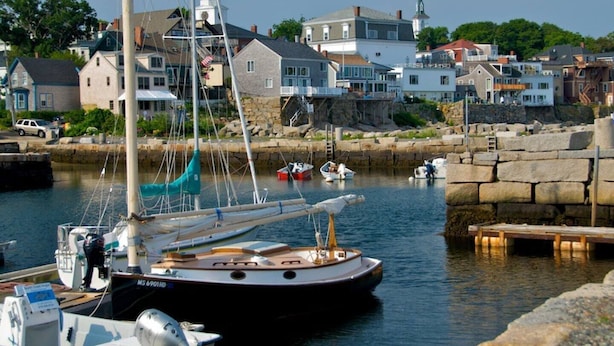 A harbor with a sailboat docked, a stone breakwater, and houses in the background.