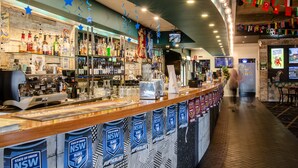 Una zona de bar con una barra que exhibe diferentes botellas y vasos, una máquina de café y una pared decorada con banderas y estandartes.