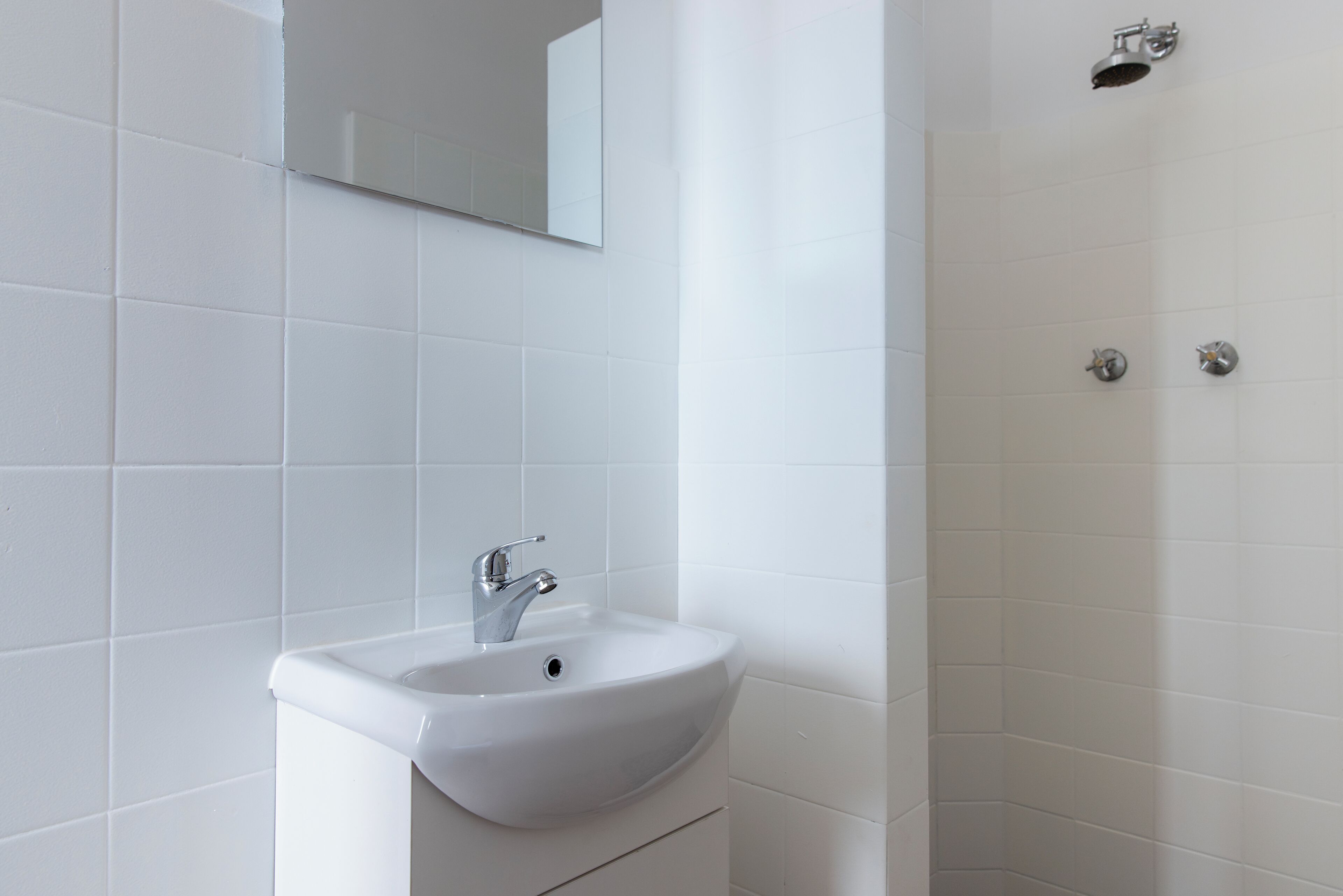 A bathroom with a white sink, a mirror above it, and a shower area with a showerhead and two controls.