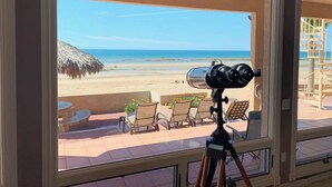 A telescope on a balcony overlooking a beach with lounge chairs and a thatched umbrella.