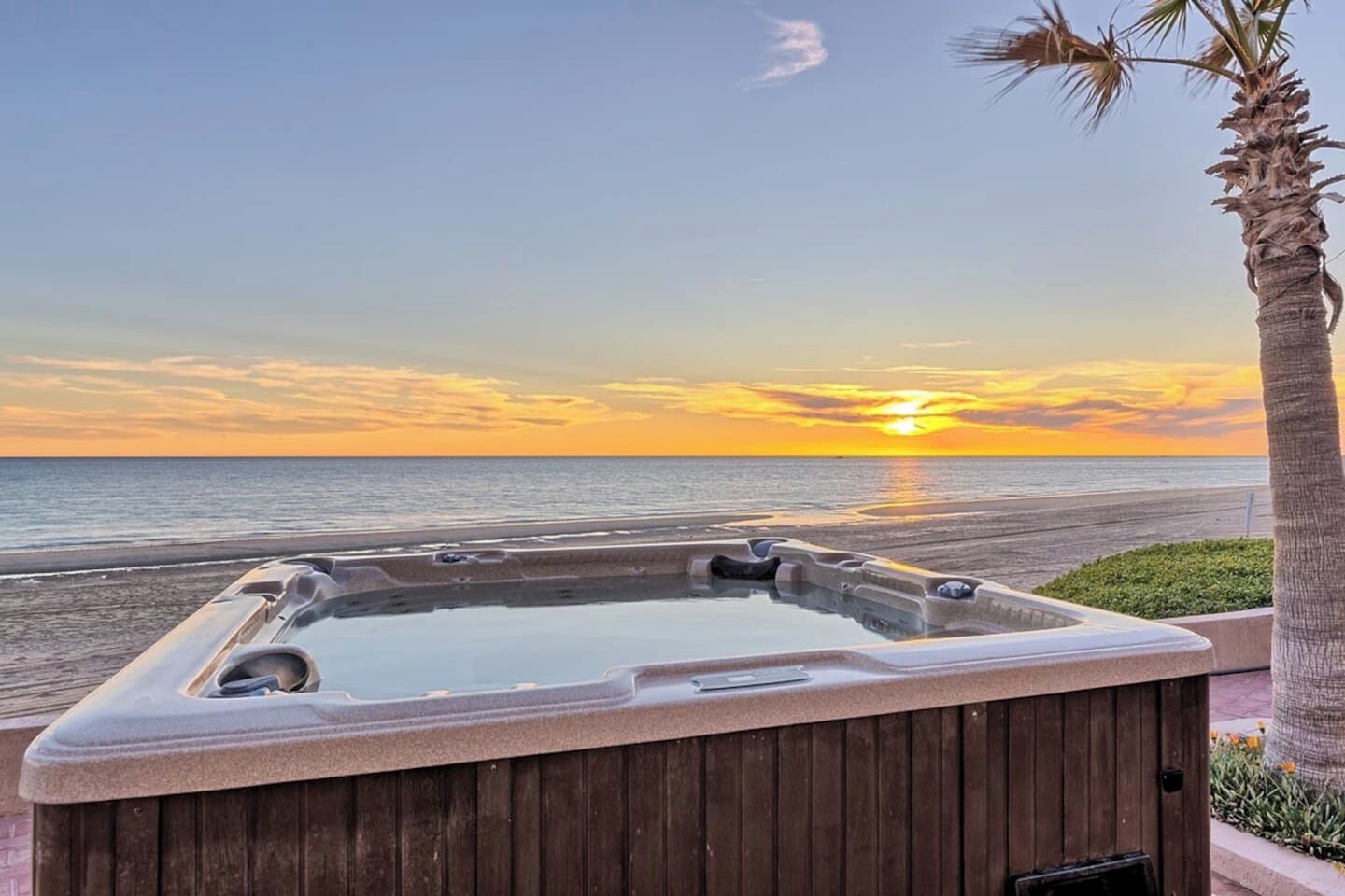 A hot tub with a wooden exterior, positioned on a wooden deck with a view of the ocean and a sunset in the background.