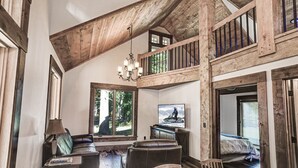 A rustic interior with a wooden ceiling, a chandelier, a television, and a view of the outdoors through large windows.