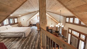 A loft bedroom with a sloped ceiling, wooden beams, a bed, a desk, and a view of the outside through a window.