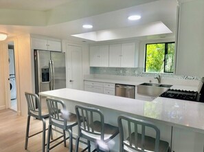 A modern kitchen with white cabinets, a stainless steel refrigerator, a built-in oven, and a large island with bar stools. - 4 Bdrm Private Jungle House, minutes to all, patios, fire pit,  parking, trees! (San Diego)