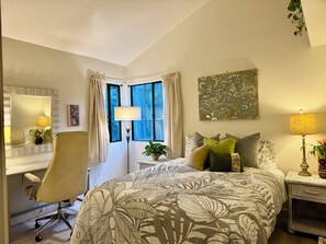 A hotel room with a bed, a desk with a chair, a window with curtains, and a wall-mounted lamp.