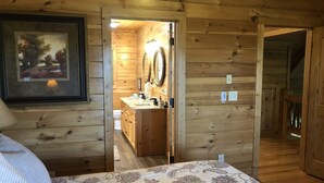 A wooden cabin interior with a bed, a bathroom with a sink and mirrors, and a framed picture on the wall. - Cozy Getaway Long Range View Paved Access Minutes To Downtown Blue Ridge Hot Tub (Blue Ridge)