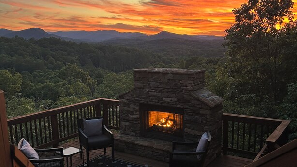 A deck with a stone fireplace, outdoor furniture, and a scenic view of a sunset over a forested landscape. - Cozy Getaway Long Range View Paved Access Minutes To Downtown Blue Ridge Hot Tub (Blue Ridge)