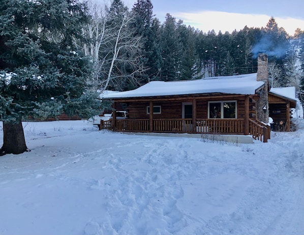 A wooden cabin with a porch, surrounded by snow-covered ground and trees.