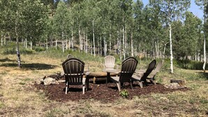 Een bebost gebied met een houten tafel en vier Adirondack-stoelen.