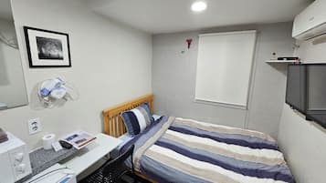 A small, neatly arranged bedroom with a single bed, a bedside table with a lamp, a framed picture on the wall, and a window with a blind.