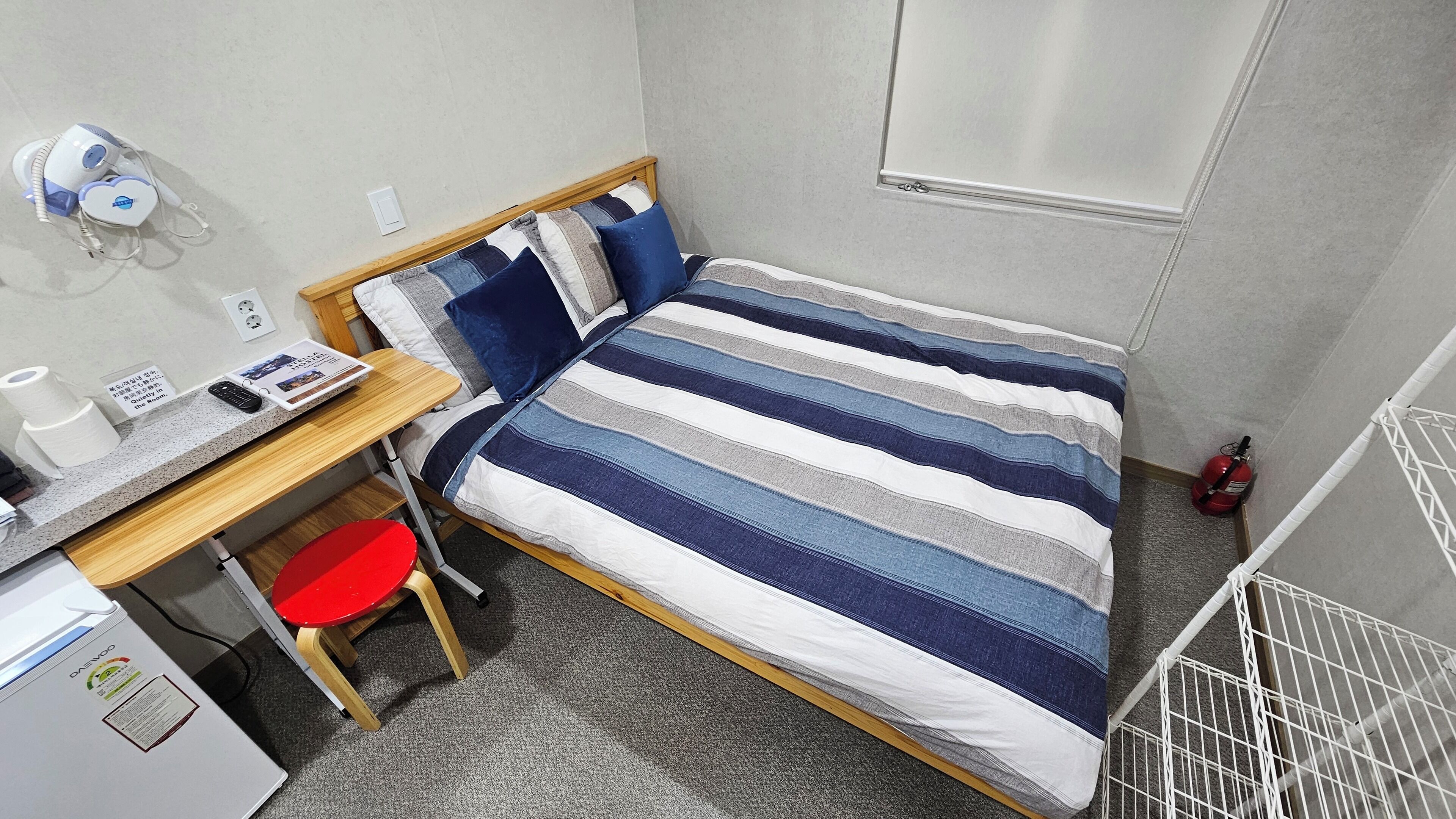 Een kleine hotelkamer met een eenpersoonsbed, een bureau met computer, een rode stoel en een kleine kitchenette.