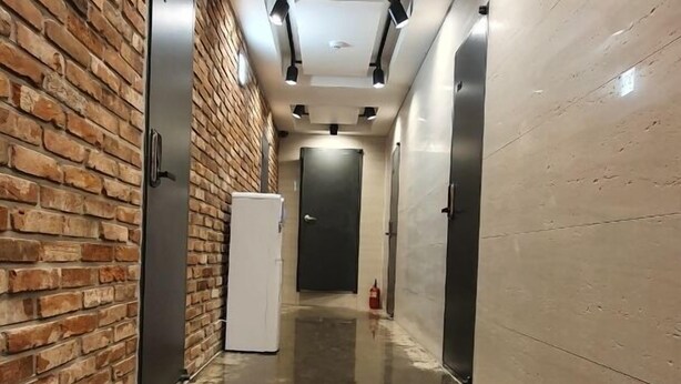Ein Flur mit poliertem Betonboden, einer Backsteinwand auf der einen Seite und einer schlichten Wand auf der anderen. Mehrere Türen und ein Feuerlöscher sind zu sehen.