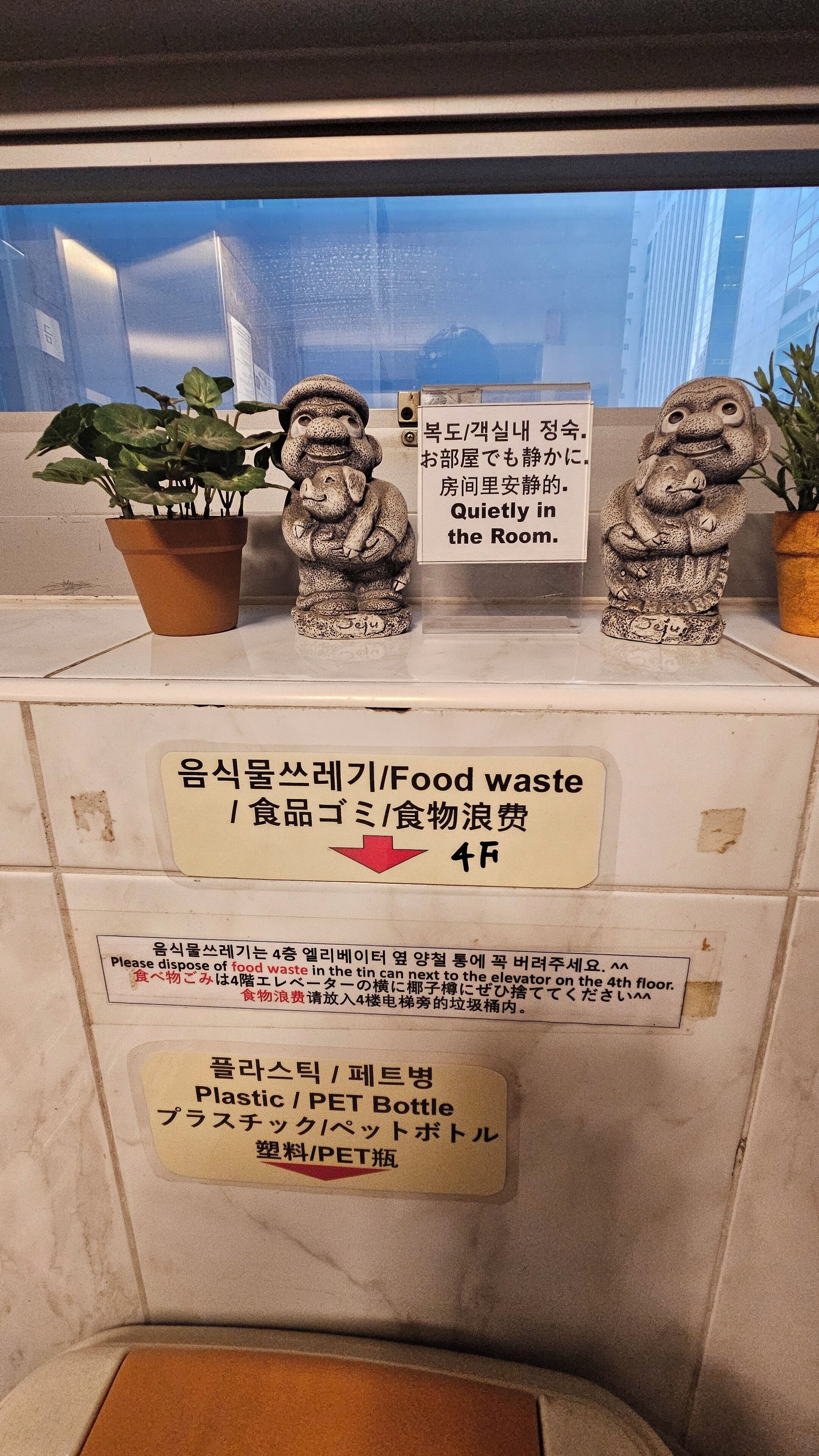 A window with a view of a building, two stone statues, a potted plant, and signs in Korean about quietness and waste disposal.