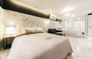 Une chambre d’hôtel avec un grand lit, une table de chevet, une lampe, une chaise et une salle de bain visible à travers une paroi en verre.