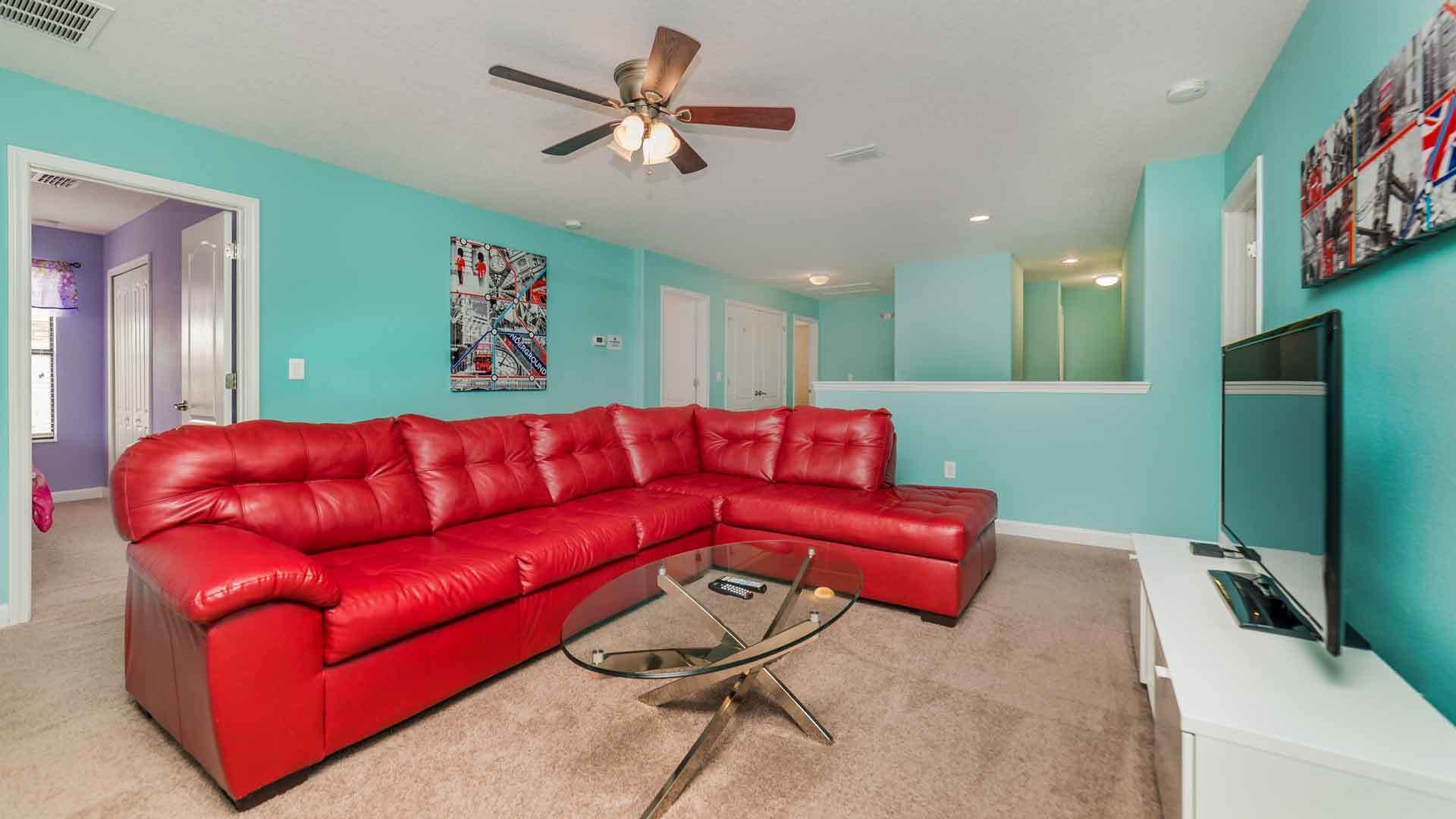 Una sala de estar con un sofá de cuero rojo, una mesa de centro de vidrio, un ventilador de techo y un televisor montado en la pared.