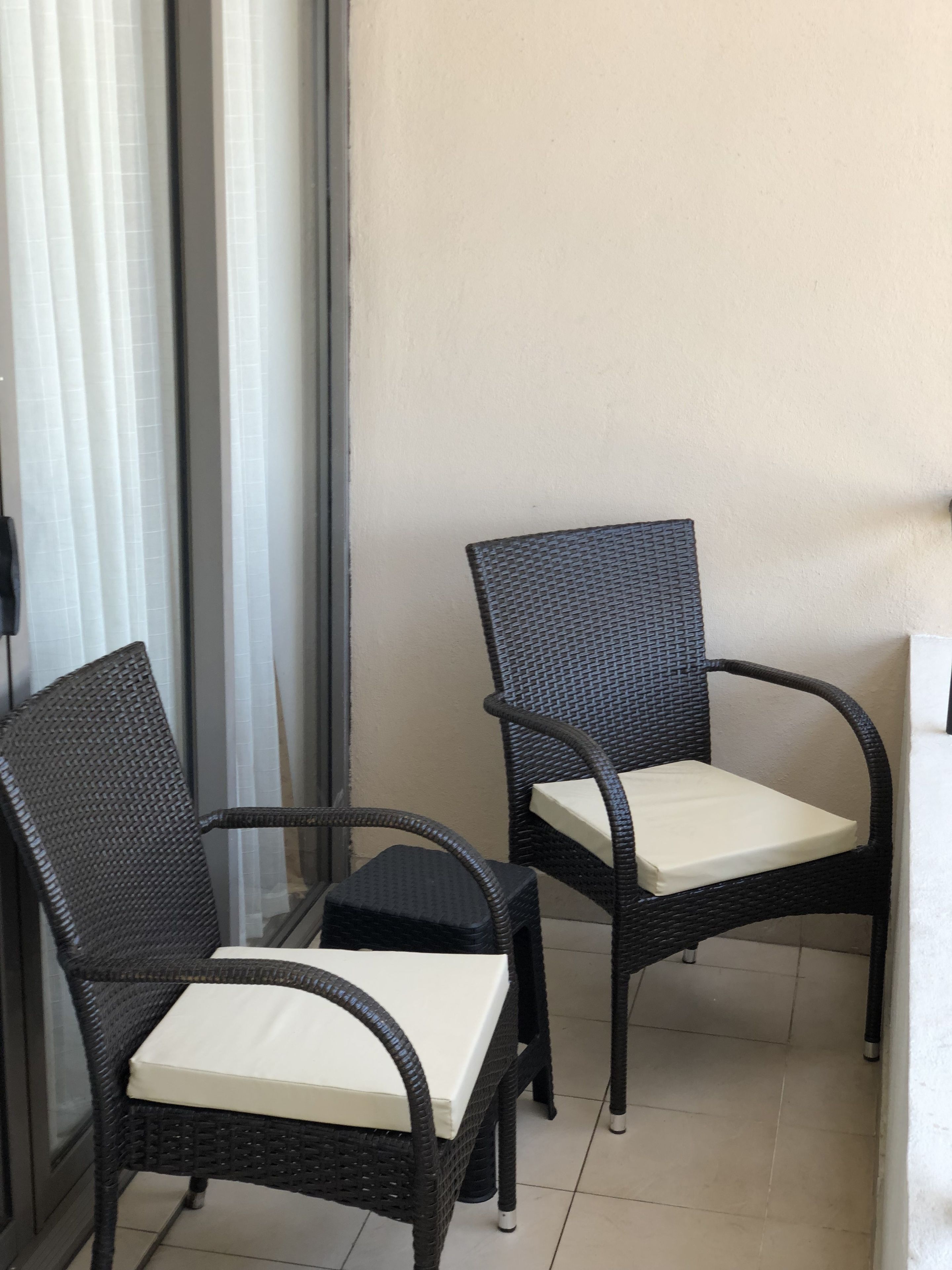 Two wicker chairs with white cushions on a tiled floor, positioned near a window with white curtains.