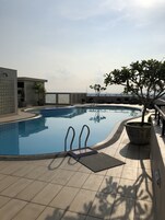 Una piscina en la azotea con agua cristalina, rodeada de plantas en macetas y un edificio moderno con grandes ventanales.