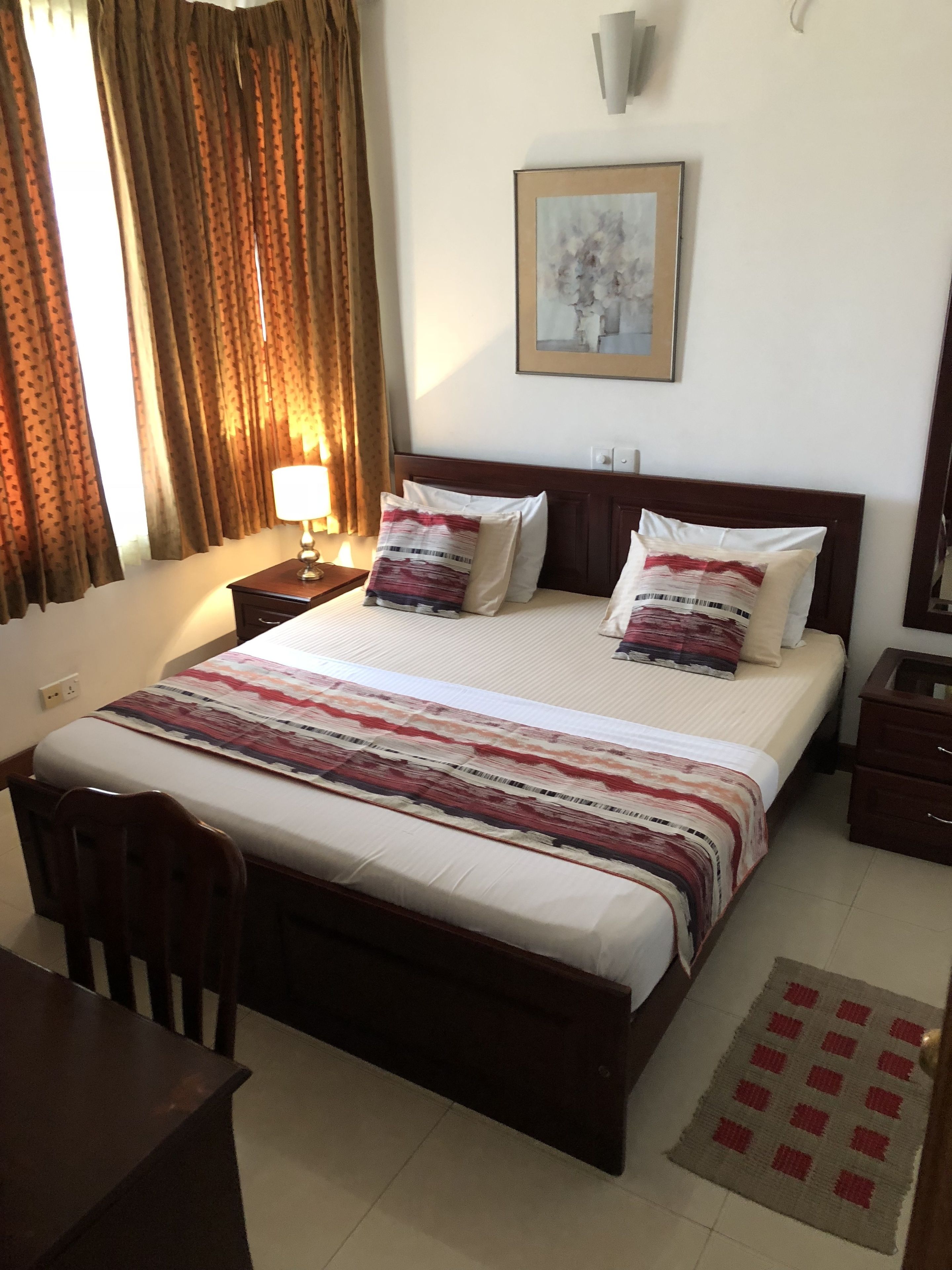 Habitación de hotel ordenada con una cama, una silla, una mesita de noche, una lámpara y un cuadro en la pared.