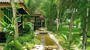 Un jardín exuberante con un elemento acuático, una estructura de techo de paja y un sendero que atraviesa la vegetación.