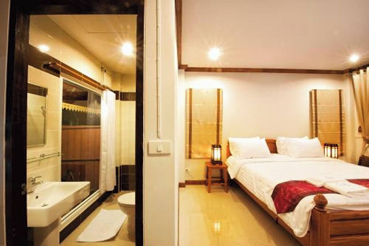 A hotel room with a wooden bed, a bedside table with a lamp, a large mirror, and a bathroom with a sink and toilet.