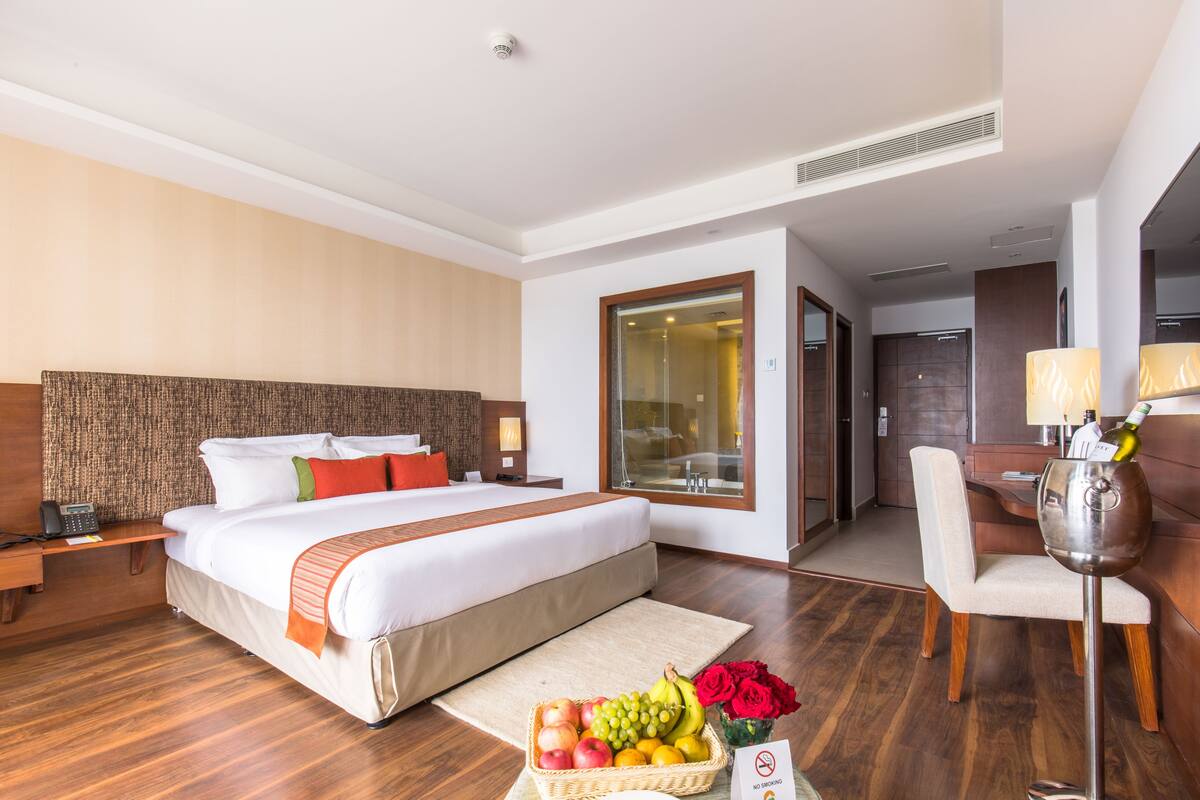 Habitación de hotel con una cama grande, una mesita de noche con un teléfono, una cesta de fruta, un jarrón de flores y una lámpara.