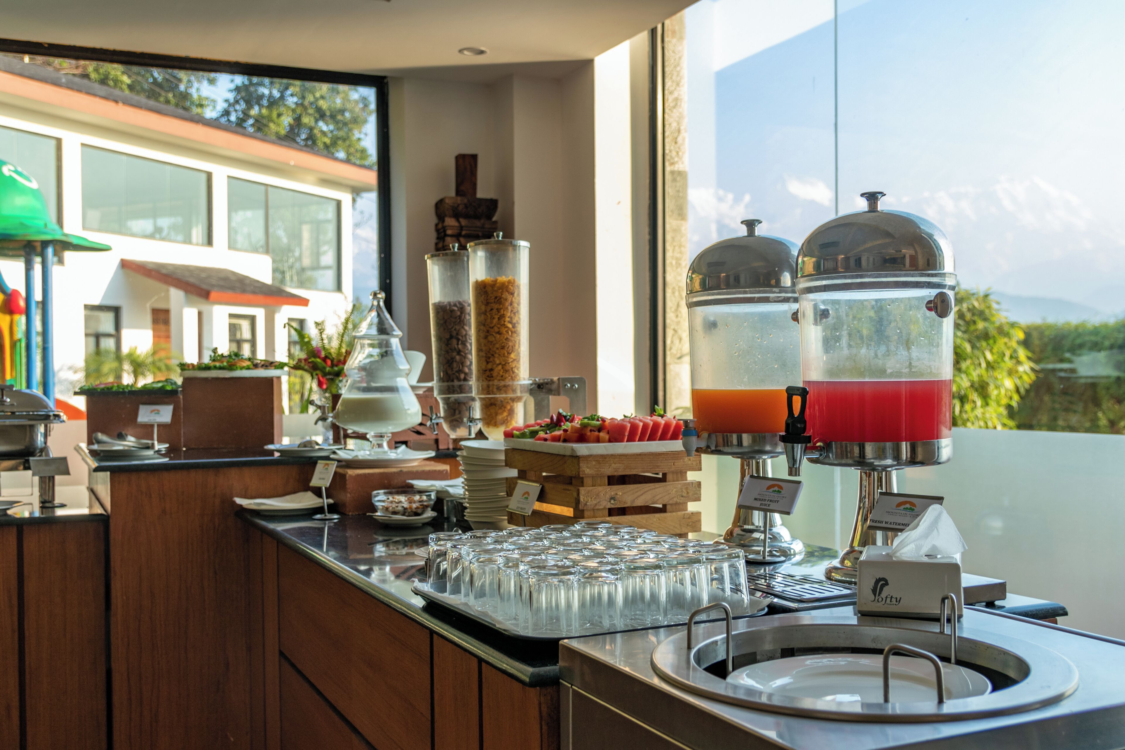 A buffet setup with a variety of food items, a juice dispenser, and a view of a building through the window.