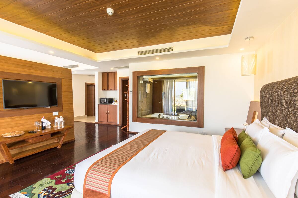Habitación de hotel con una cama grande, un televisor de pantalla plana, un mueble de entretenimiento de madera y un baño con un espejo grande y bañera.
