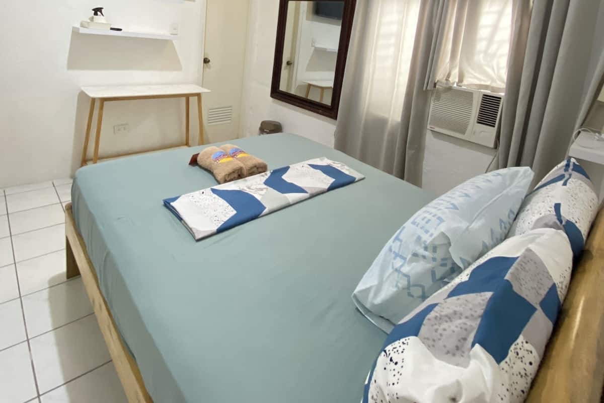 Een slaapkamer met een bed, een aan de muur bevestigde televisie, een raam met gordijnen en een klein plankje met toiletartikelen.