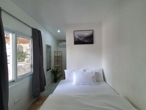 Een slaapkamer met een bed, een raam met gordijnen, een aan de muur hangend schilderij en een kleine plant.