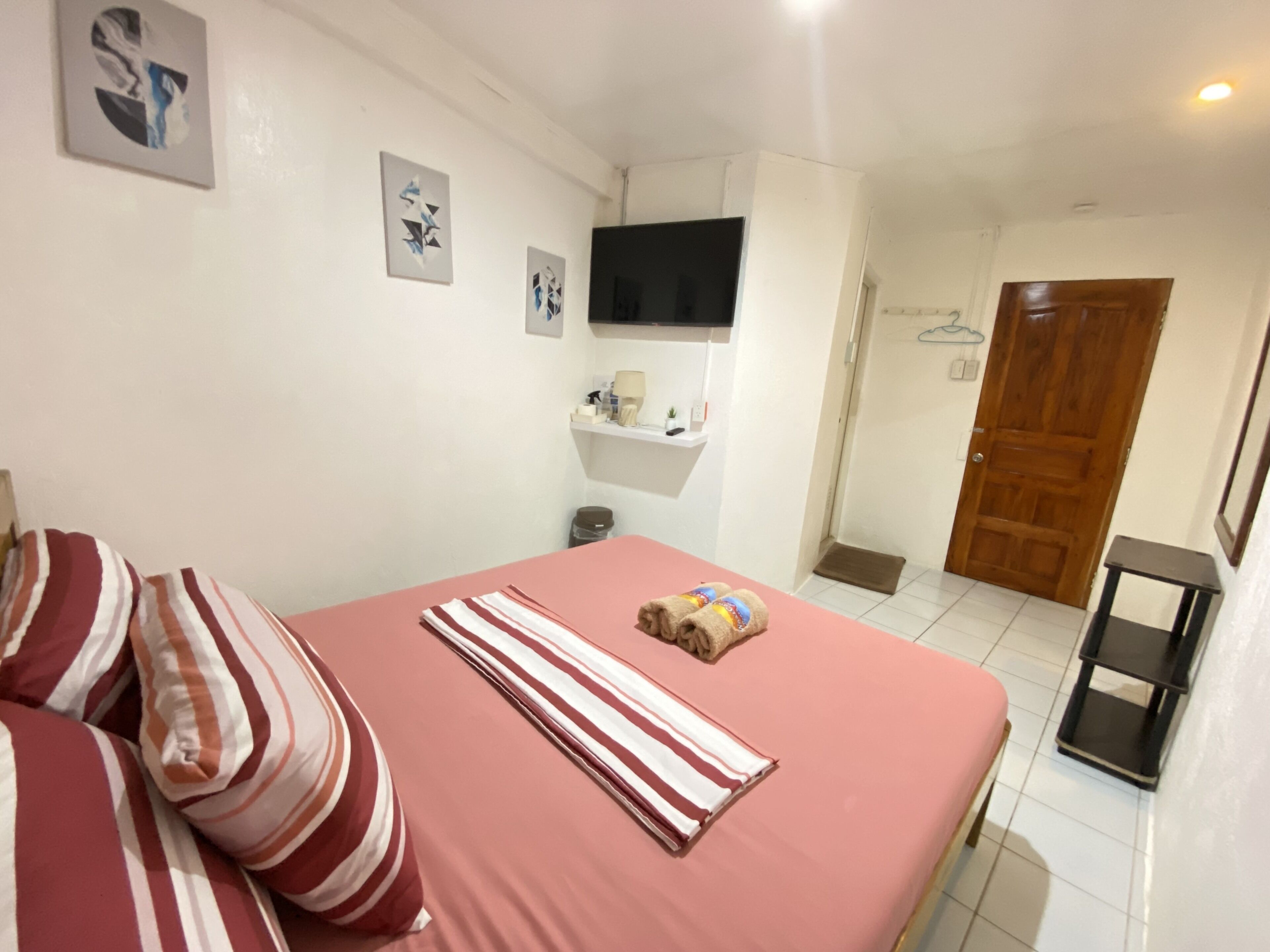 A bedroom with a bed, pillows, a TV mounted on the wall, a shelf with toiletries, and a wooden door.