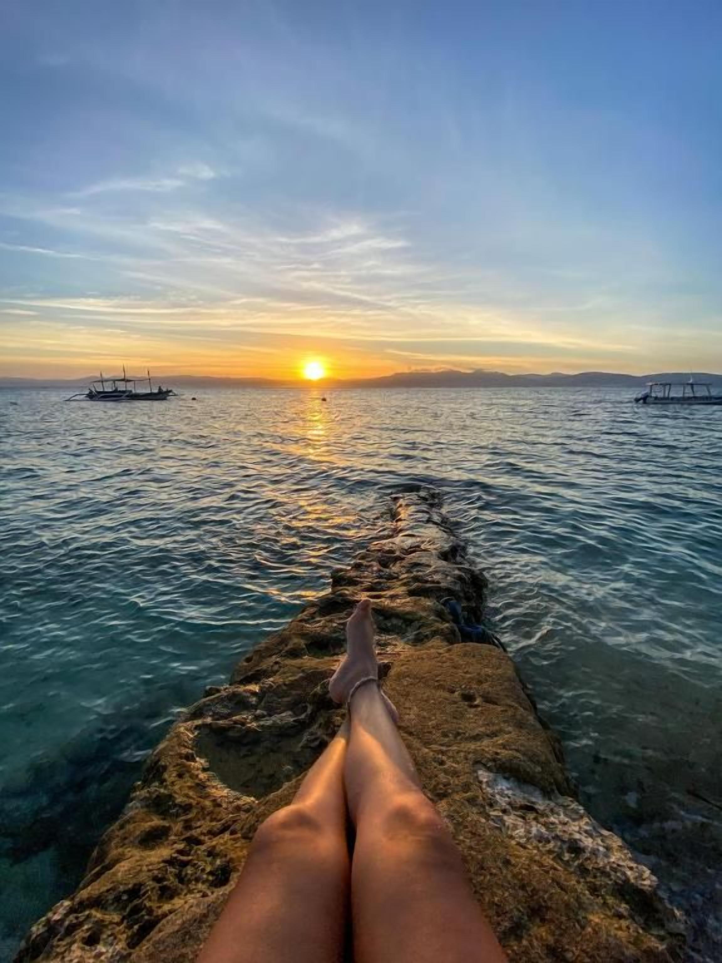 A person sitting on a rocky ledge overlooking a body of water with boats and a sunset in the distance.