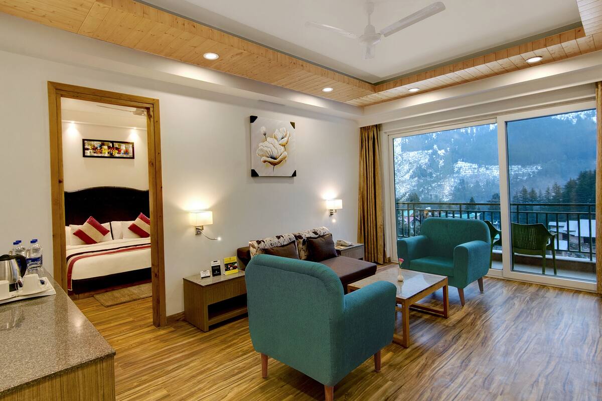 Habitación de hotel con cama, sofá, sillón, mesa y balcón con vista a la montaña.