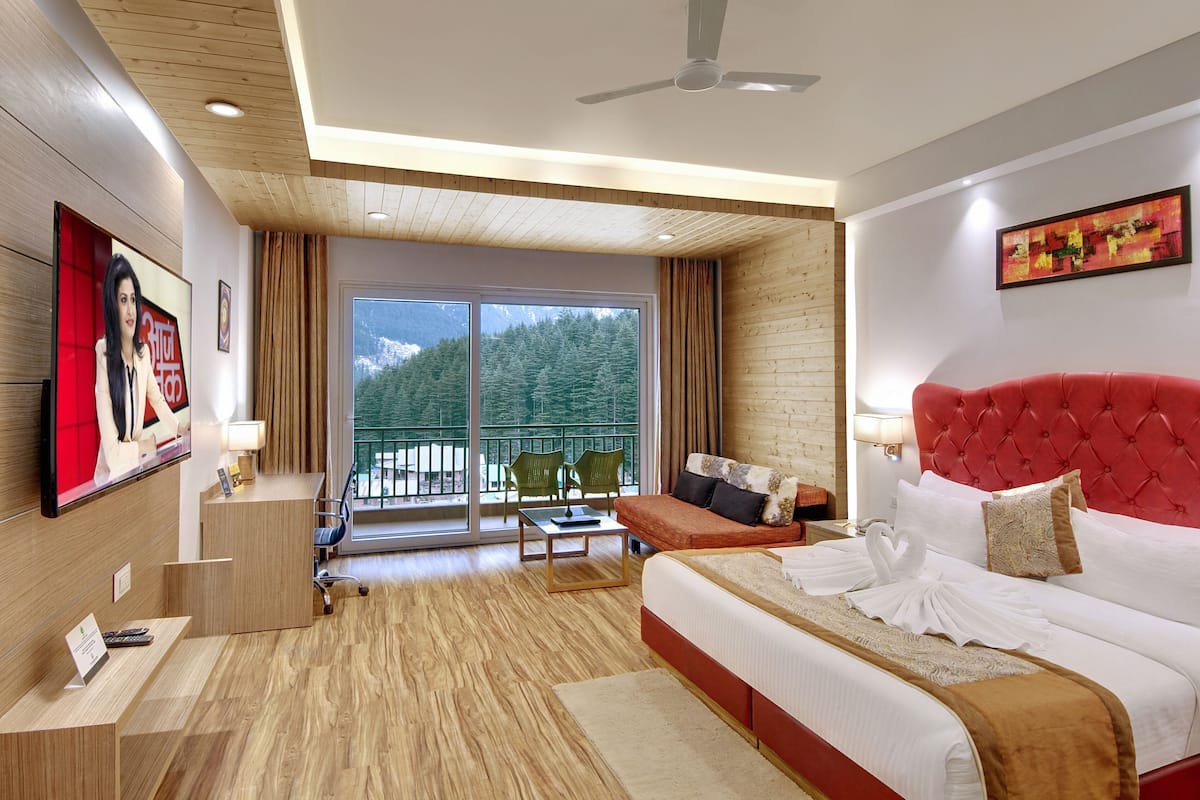 Habitación de hotel con cama, escritorio, silla, balcón y ventilador de techo, con vistas a árboles.
