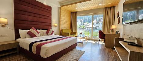 A modern hotel room with a large bed, wooden flooring, a desk, and a balcony with a view of buildings.