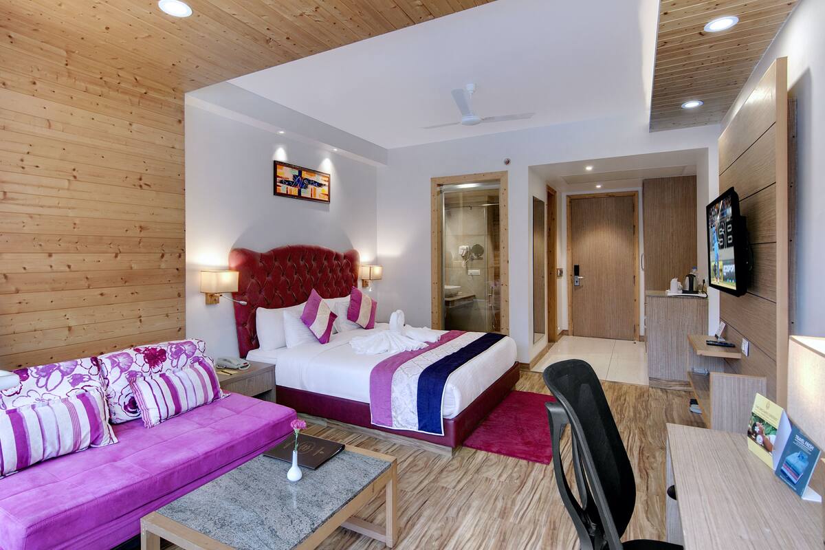 Habitación de hotel con cama, sofá, escritorio, televisor y baño visible a través de una puerta abierta.