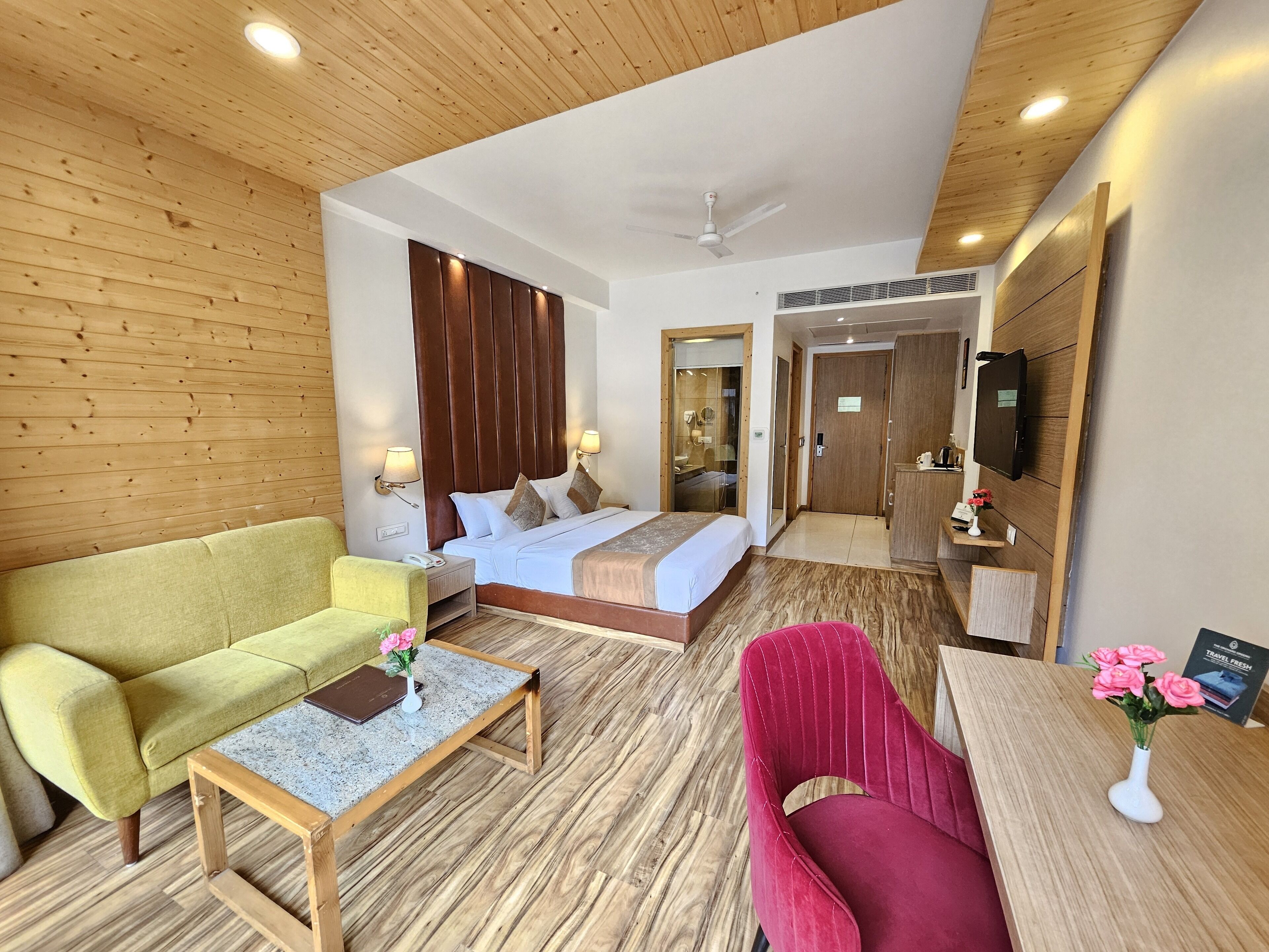 A hotel room with a bed, a green sofa, a red chair, a small table with a vase of flowers, and a wooden floor.