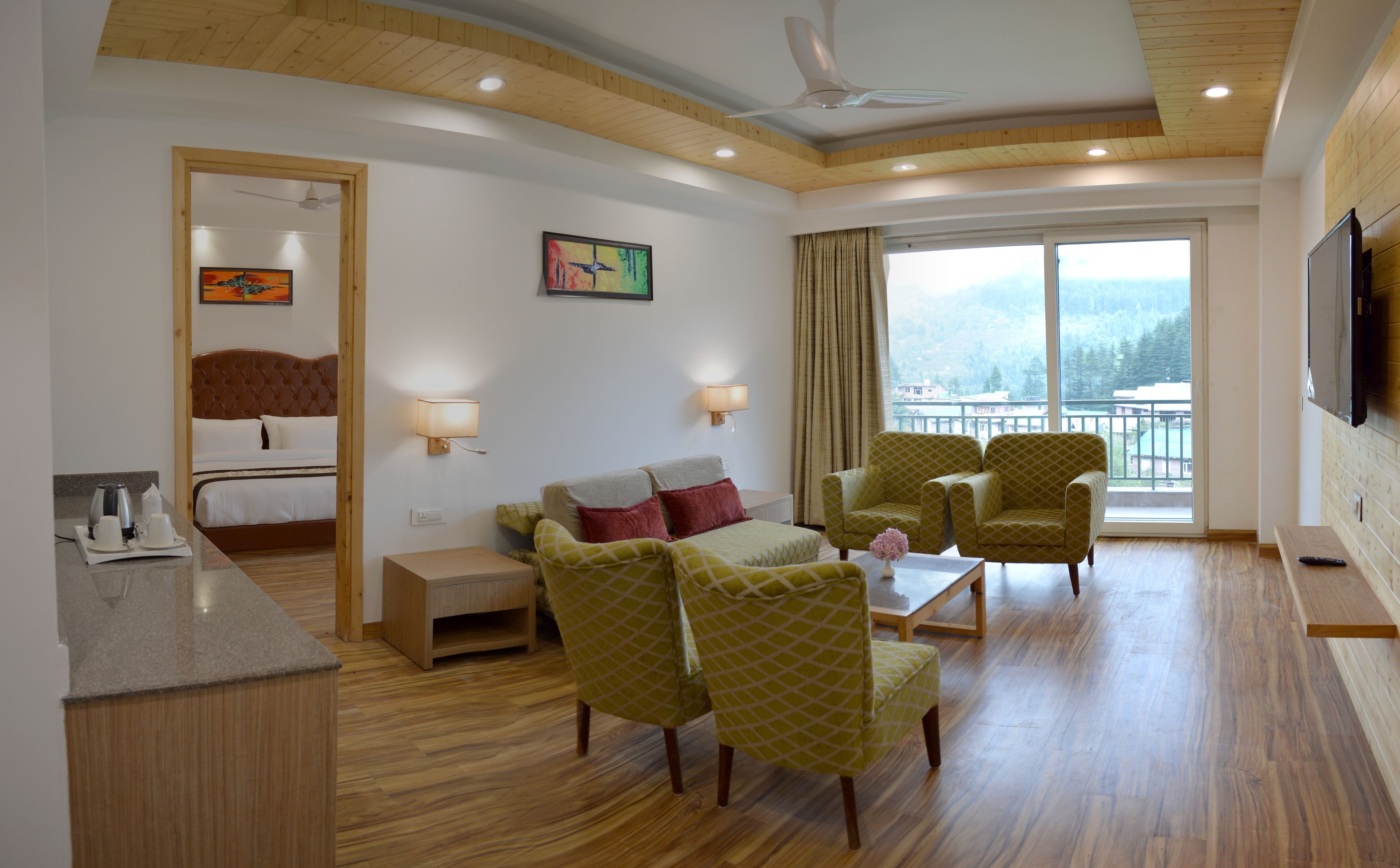 Habitación de hotel con cama, sofá, dos sillones, una mesa de centro, televisor y balcón con vistas.