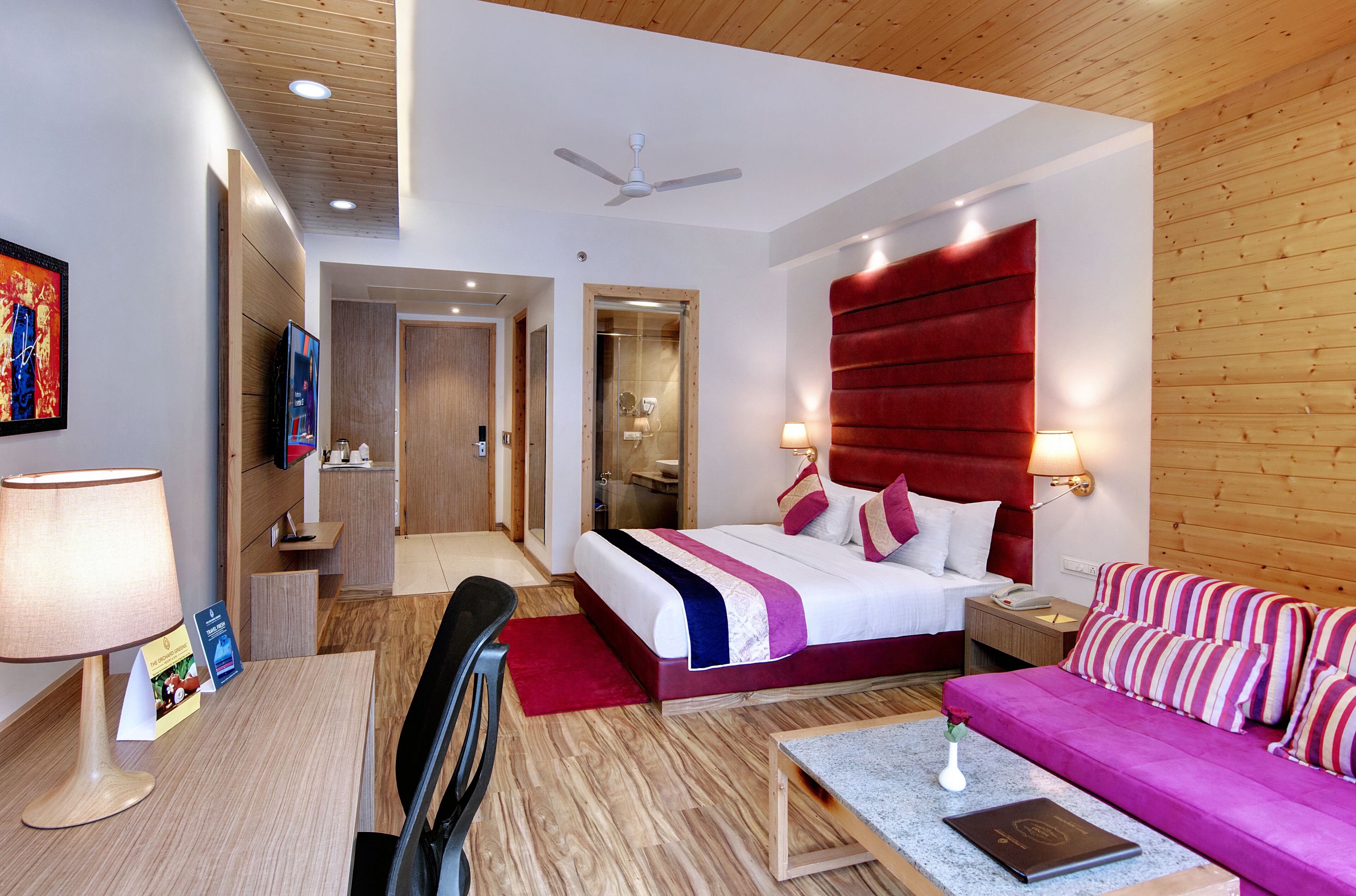 A modern hotel room with a large bed, a red headboard, a sofa, a desk with a lamp, and a television.