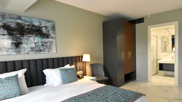 A modern hotel room with a large bed, a bedside table with a lamp, a chair, and a bathroom visible through an open door. - Capella Otel (Eskisehir)