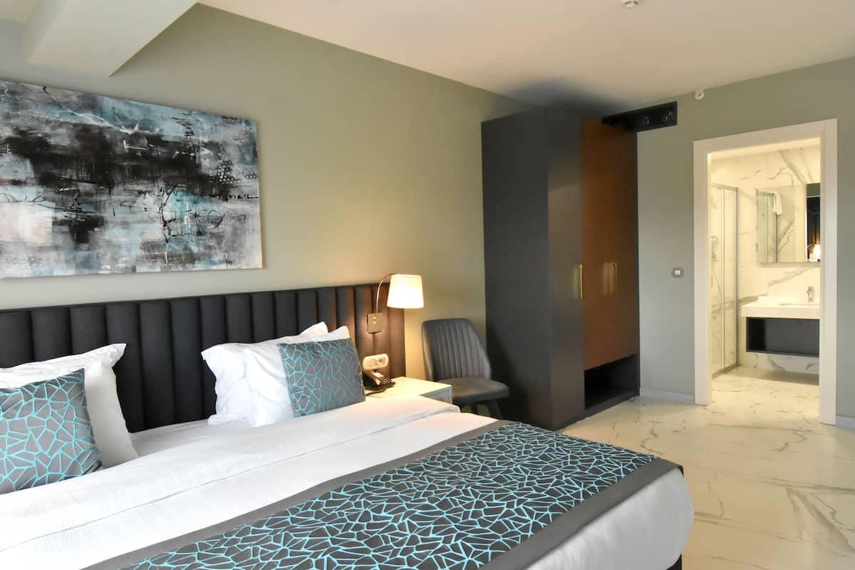 A modern hotel room with a large bed, a bedside table with a lamp, a chair, and a bathroom visible through an open door.