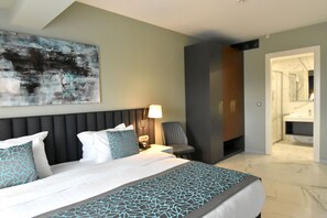 A modern hotel room with a large bed, a bedside table with a lamp, a chair, and a bathroom visible through an open door.