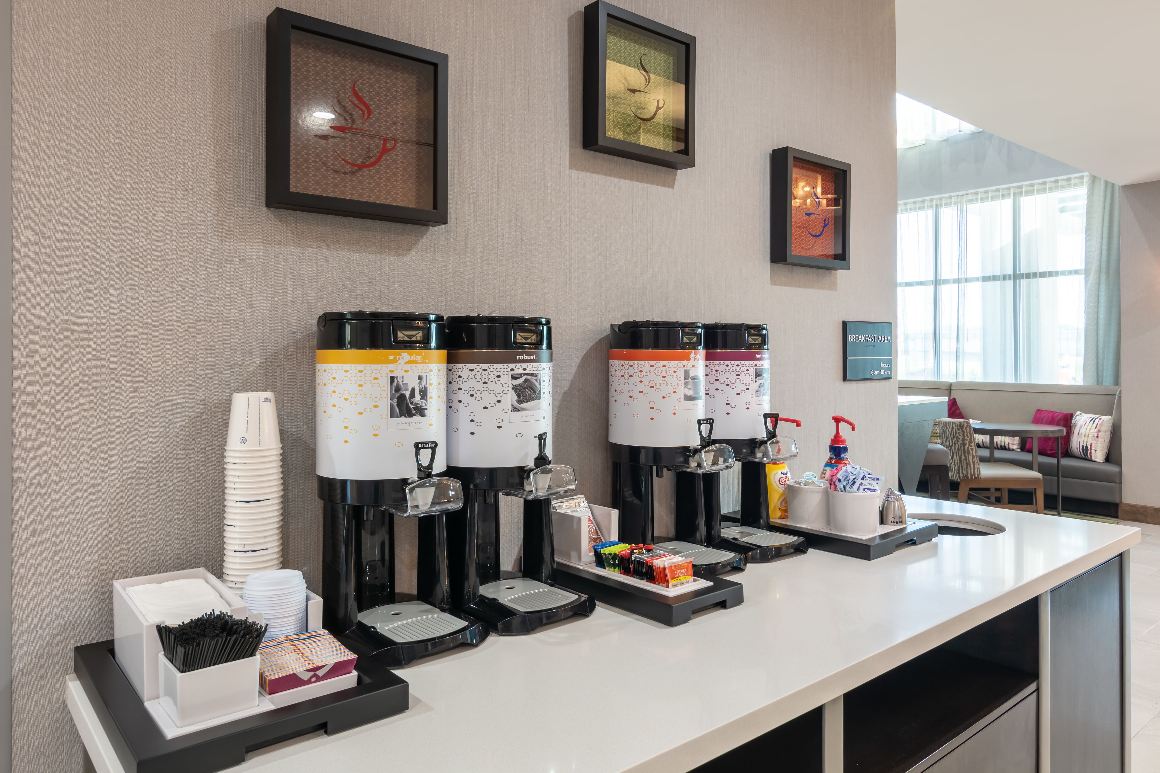 A breakfast bar with multiple coffee dispensers, a stack of white cups, and a tray with condiments.
